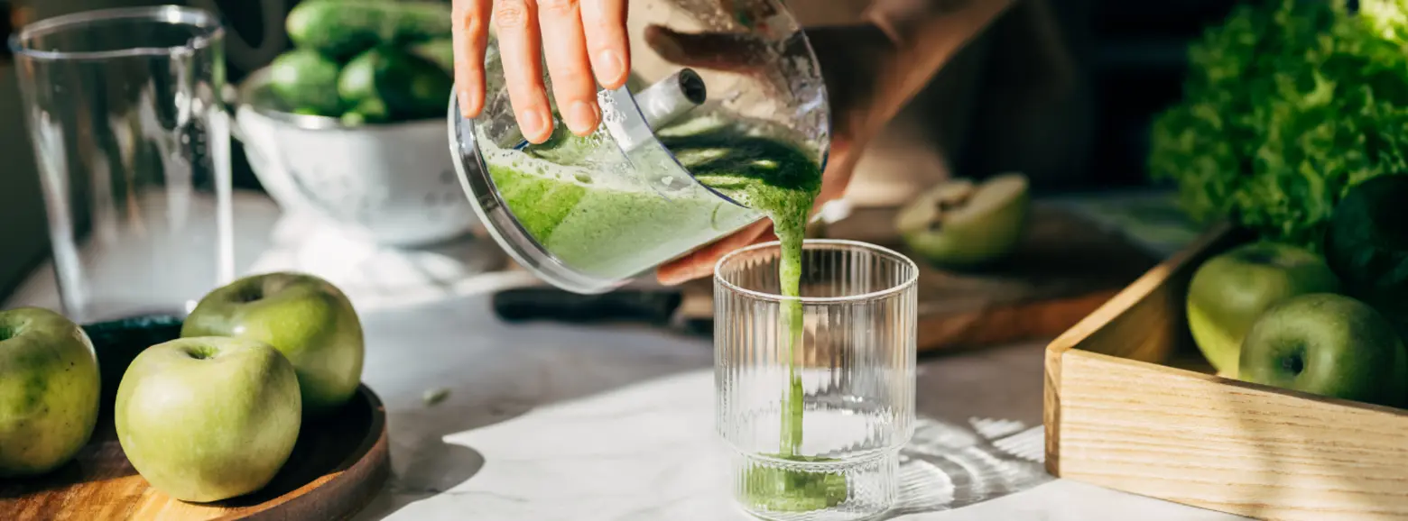 A hand pours a green smoothie from a blender into a clear glass on a countertop. Green apples and cucumbers are visible nearby, emphasizing a fresh and healthy vibe in the kitchen setting.