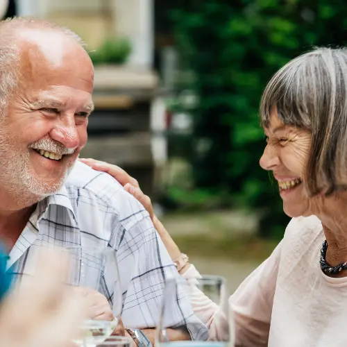 A joyful elderly couple shares a moment at a gathering, smiling and laughing with each other. The man, with a white beard and a striped shirt, touches the woman's shoulder. She has gray hair and wears a light-colored top, radiating warmth and connection.