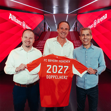 Na slici su tri muškarca koji drže dres fudbalskog kluba FC Bayern München. Dres je narandžaste boje, sa natpisom "2027 DOPPELHERZ" na poleđini. Iza njih se nalazi hodnik sa natpisom "Allianz Arena" u pozadini.