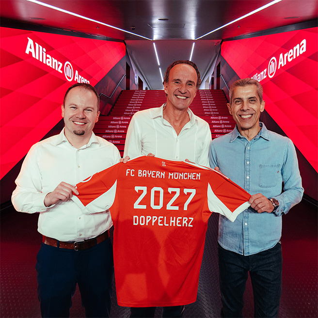 Na slici su tri muškarca koji drže dres fudbalskog kluba FC Bayern München. Dres je narandžaste boje, sa natpisom "2027 DOPPELHERZ" na poleđini. Iza njih se nalazi hodnik sa natpisom "Allianz Arena" u pozadini.